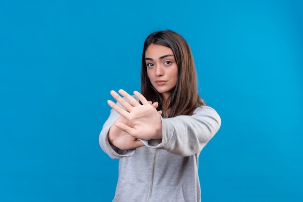 Junges schönes Mädchen im grauen Kapuzenpulli, das Kamera betrachtet und Stoppgeste macht, die über blauem Hintergrund steht