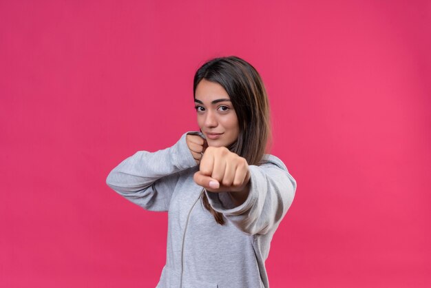 Junges schönes Mädchen im grauen Kapuzenpulli, der die geballte Faust zur Kamera mit dem Lächeln auf Gesicht betrachtet, das über rosa Hintergrund steht