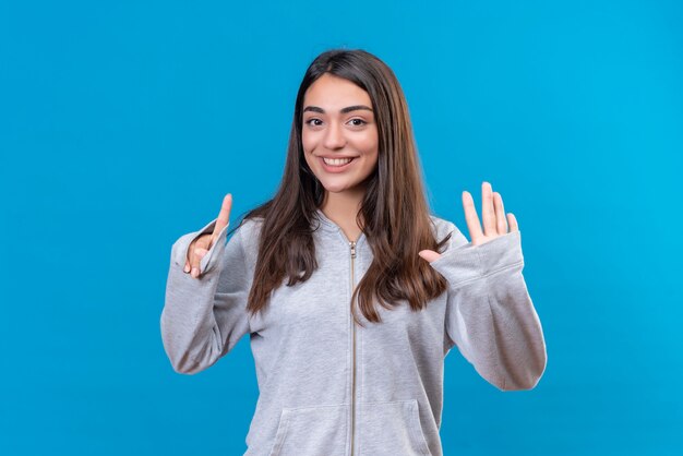 Junges schönes Mädchen im grauen Kapuzenpulli, der Kamera mit Lächeln auf Gesicht betrachtet, das eine und fünf Zahlen auf Hand steht, die über blauem Hintergrund stehen