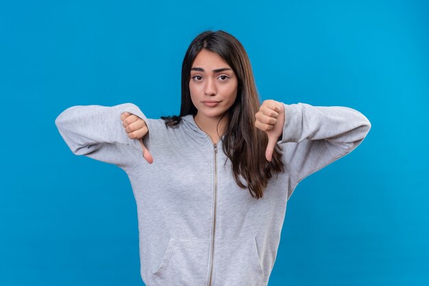 Junges schönes Mädchen im grauen Kapuzenpulli, der Kamera mit unzufriedenem Gesicht betrachtet, das Abneigungsgeste macht, die über blauem Hintergrund steht