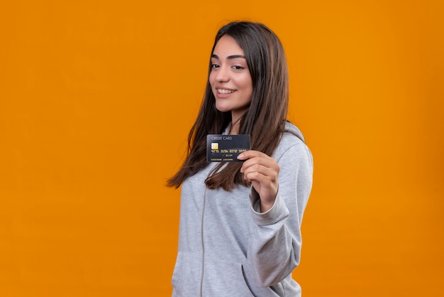Junges schönes Mädchen im grauen Kapuzenpulli, der Kreditkarte hält und Kreditkarte mit Lächeln auf Gesicht steht, das über orange Hintergrund steht