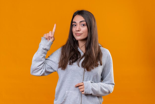 Junges schönes Mädchen im grauen Kapuzenpulli, der nach oben zeigt, die große Idee hat, zuversichtlich über orange Hintergrund stehend zu lächeln