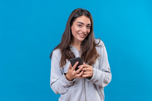 Junges schönes Mädchen im grauen Kapuzenpulli, der Telefon betrachtet Kamera mit Vergnügen steht über blauem Hintergrund