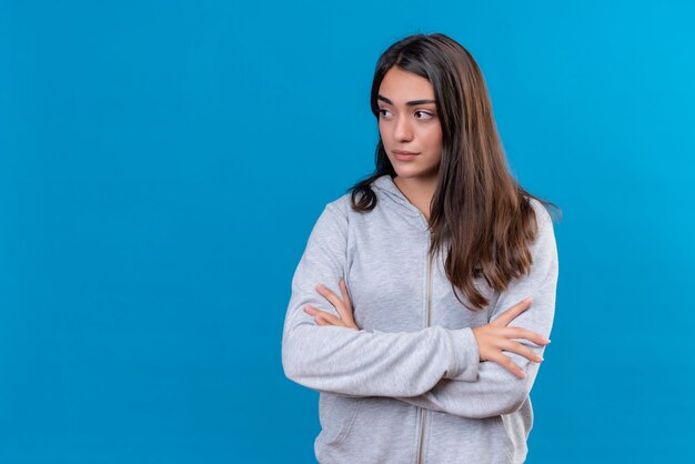 Junges schönes Mädchen im grauen Kapuzenpulli, der weg mit ärgerlicher Emotion steht, die über blauem Hintergrund steht