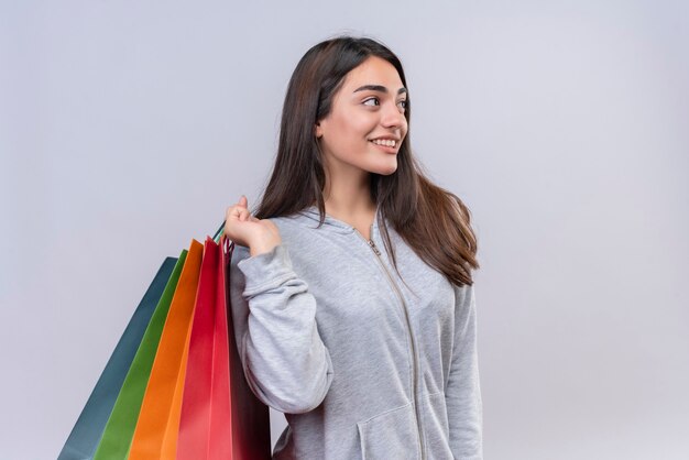 Junges schönes Mädchen im grauen Kapuzenpulli, der weg mit Lächeln auf Gesicht hält, das Pakete hält, die über weißem Hintergrund stehen