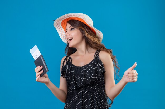 Junges schönes Mädchen im Kleid im Tupfen im Sommerhut, der Flugtickets lächelnd beiseite schauend zeigt Daumen hoch stehend über blauem Hintergrund