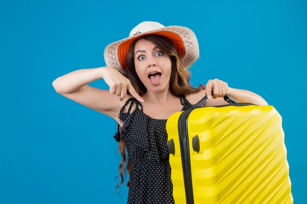 Junges schönes Mädchen im Kleid im Tupfen im Sommerhut, der Koffer hält, der mit dem Finger auf ihn schaut und Kamera überrascht über blauem Hintergrund steht