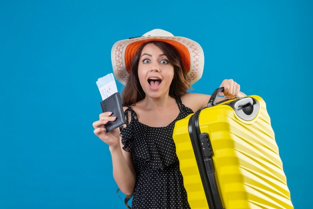 Junges schönes Mädchen im Kleid im Tupfen im Sommerhut stehend mit Koffer, der Flugtickets hält, die über blauem Hintergrund überrascht und glücklich schauen