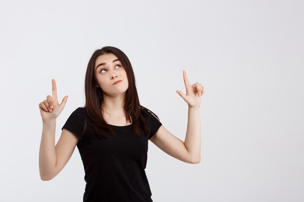 Junges schönes Mädchen im schwarzen T-Shirt, das Finger oben über weißer Wand zeigt