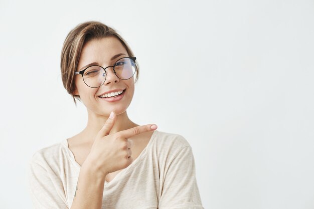 Junges schönes Mädchen in den Gläsern, die lächelnden Zeigefinger in der Seite lächeln.