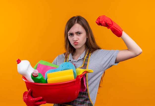 Junges schönes Mädchen in der Schürze und in den Gummihandschuhen, die Becken mit Reinigungswerkzeugen halten, die Faust heben, die langlebig aussieht, sel zufrieden, bereit zu reinigen