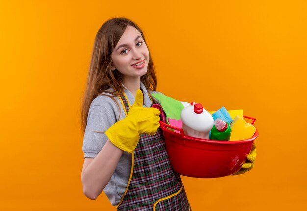 Junges schönes Mädchen in der Schürze und in den Gummihandschuhen, die Becken mit Reinigungswerkzeugen halten, die fröhlich lächelnd Daumen zeigen