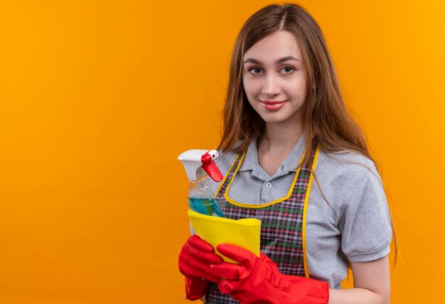 Junges schönes Mädchen in der Schürze und in den Gummihandschuhen, die Reinigungsspray und Teppich halten, die Kamera lächelnd betrachten, bereit zum Reinigen