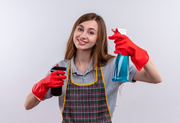 Junges schönes Mädchen in der Schürze und in den Gummihandschuhen, die Reinigungssprays halten, die fröhlich lächeln