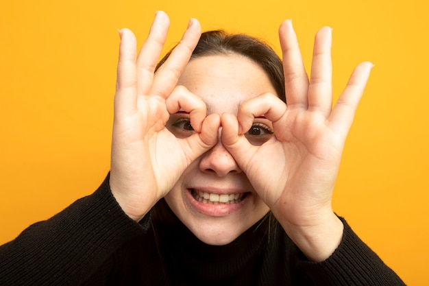 Junges schönes Mädchen in einem schwarzen Rollkragenpullover, der ok Zeichen wie Fernglas mit Fingern durch Finger macht, die fröhlich lächeln