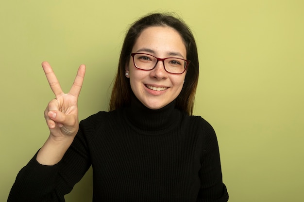 Kostenloses Foto junges schönes mädchen in einem schwarzen rollkragenpullover und in den glücklichen und positiven v-zeichen der brille