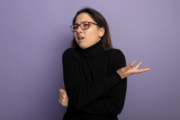 Junges schönes Mädchen in einem schwarzen Rollkragenpullover und in der Brille, die beiseite schauen, verwirrt mit dem ausgestreckten Arm als fragend