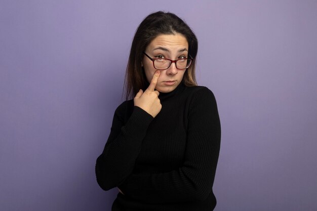 Junges schönes Mädchen in einem schwarzen Rollkragenpullover und in der Brille, die mit dem Finger auf ihr Auge zeigen, das skeptisch ist