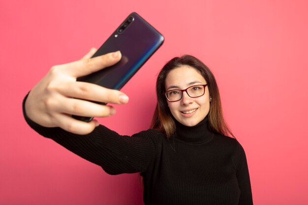 Junges schönes Mädchen in einem schwarzen Rollkragenpullover und in der Brille unter Verwendung des Smartphones, das selfie tut, das mit glücklichem Gesicht lächelt