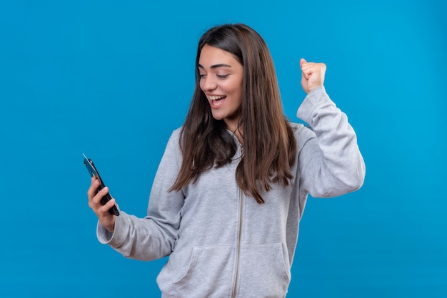 Junges schönes Mädchen in grauem Blick auf das Telefon mit Lächeln auf Gesicht und Halten des Kreditkartentelefons, das über blauem Hintergrund steht