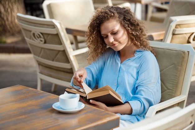 Junges schönes Mädchen lächelnd, Buch lesend, im Café sitzend.