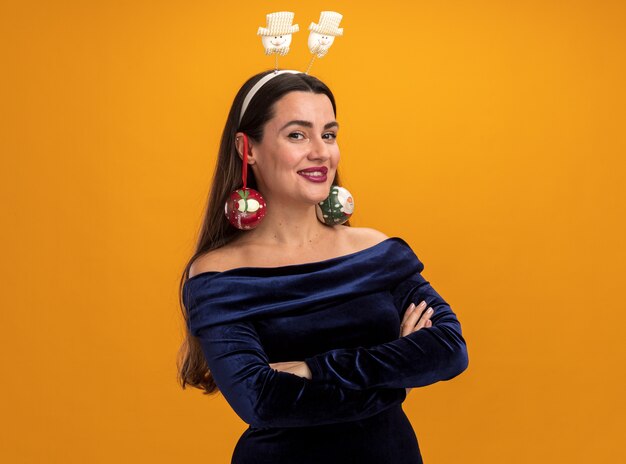 junges schönes Mädchen mit blauem Kleid und Weihnachtshaarreifen mit Weihnachtskugeln auf den Ohren - isoliert auf oranger Wand