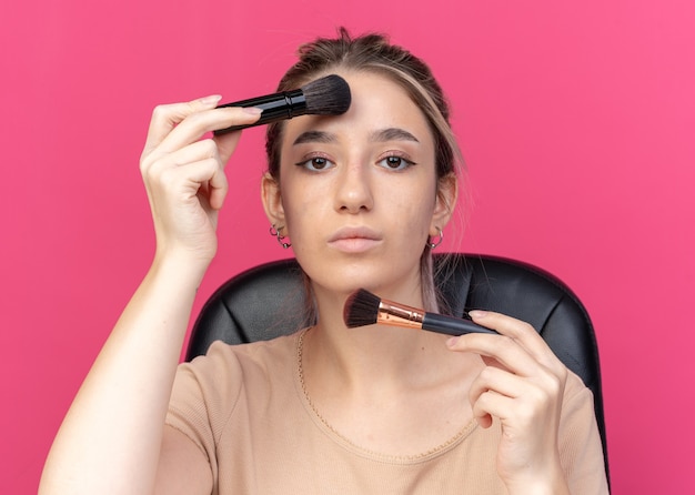 Junges schönes Mädchen mit Blick auf die Kamera sitzt am Tisch mit Make-up-Tools, die Puder erröten, isoliert auf rosa Wand