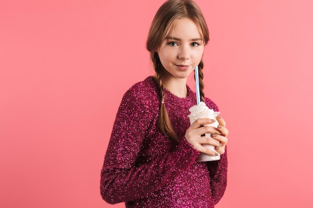 Junges schönes mädchen mit zwei zöpfen im pullover, das milchshake in den händen hält, während es glücklich in die kamera über rosa hintergrund schaut