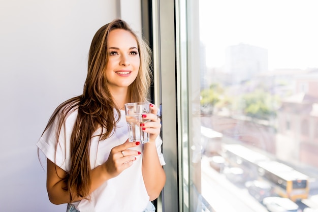 Junges schönes Mädchen oder Frau mit einem Glas Wasser nahe dem Fenster im weißen Hemd und im grauen Gewand