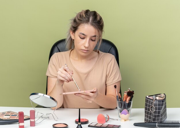 Junges schönes Mädchen sitzt am Schreibtisch mit Make-up-Tools, die Lidschatten mit Make-up-Pinsel auf olivgrünem Hintergrund auftragen