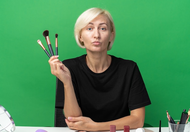 Junges schönes Mädchen sitzt am Tisch mit Make-up-Tools, die Make-up-Pinsel auf grüner Wand halten