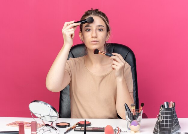 Junges schönes Mädchen sitzt am Tisch mit Make-up-Tools, die Puderrouge einzeln auf rosa Hintergrund auftragen