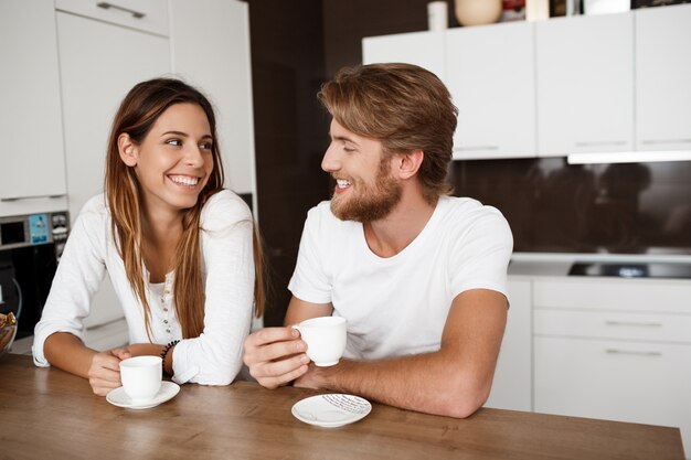 Junges schönes Paar, das am lächelnden Morgenkaffee der Küche sitzt.