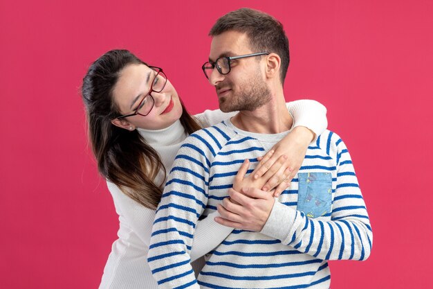 junges schönes Paar in der Freizeitkleidung glückliche Frau, die ihren lächelnden Freund glücklich in der Liebe zusammen umarmt, der Valentinstag steht über rosa Wand