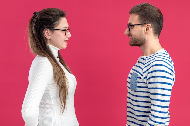 Junges schönes paar in der glücklichen kleidung des glücklichen mannes und der frau, die einander lächelnd betrachten und valentinstag feiern, der über rosa hintergrund steht