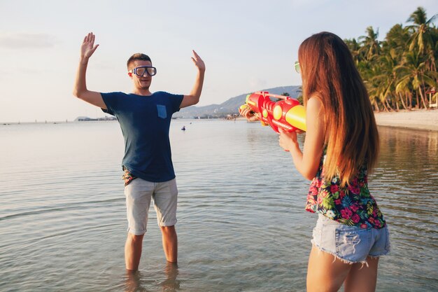 Junges schönes Paar in der Liebe, die am tropischen Strand spielt, Sommerferien, Honigmond, Romantik, Sonnenuntergang, glücklich, Spaß haben, Wasserpistole, Kampf, Mann gibt auf, positiv, lustig