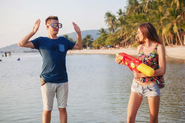 Junges schönes Paar in der Liebe, die am tropischen Strand spielt, Sommerferien, Honigmond, Romantik, Sonnenuntergang, glücklich, Spaß haben, Wasserpistole, Kampf, Mann gibt auf, positiv, lustig