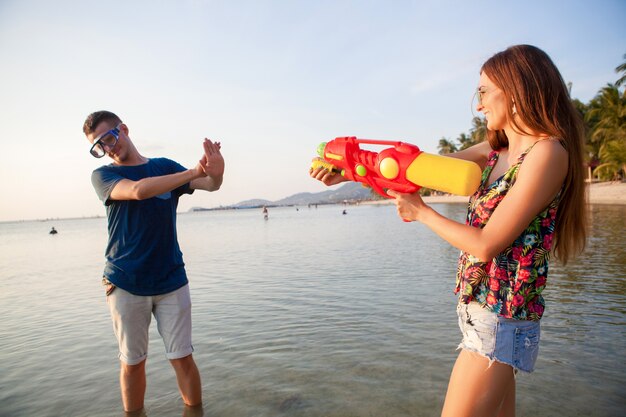 Junges schönes Paar in der Liebe, die am tropischen Strand spielt, Sommerferien, Honigmond, Romantik, Sonnenuntergang, glücklich, Spaß haben, Wasserpistole, Kampf, Mann gibt auf, positiv, lustig