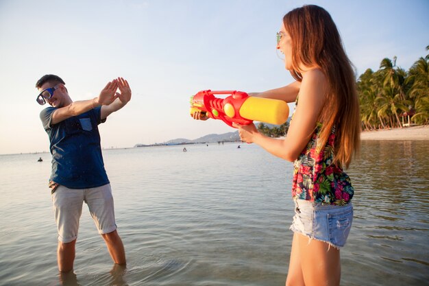 Junges schönes Paar in der Liebe, die am tropischen Strand spielt, Sommerferien, Honigmond, Romantik, Sonnenuntergang, glücklich, Spaß haben, Wasserpistole, Kampf, Mann gibt auf, positiv, lustig