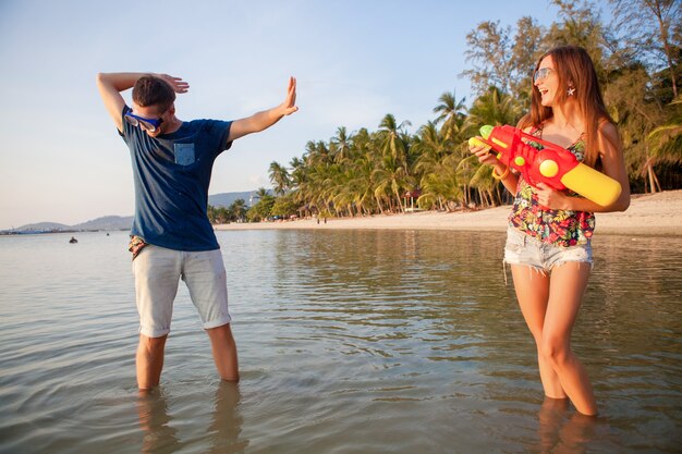 Junges schönes Paar in der Liebe, die am tropischen Strand spielt, Sommerferien, Honigmond, Romantik, Sonnenuntergang, glücklich, Spaß haben, Wasserpistole, Kampf, Mann gibt auf, positiv, lustig