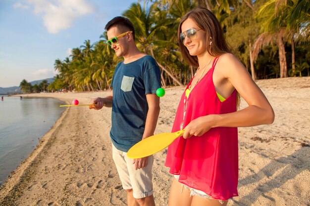 Junges schönes Paar in der Liebe, die Tischtennis am tropischen Strand spielt, Spaß, Sommerferien, aktiv, lächelnd, lustig, positiv hat