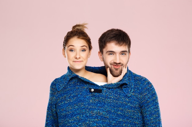Junges schönes Paar in einem blauen Strickpullover posierend lächelnd, der Spaß über hellrosa Wand hat