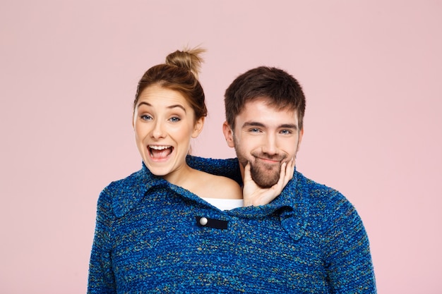 Junges schönes Paar in einem blauen Strickpullover posierend lächelnd, der Spaß über hellrosa Wand hat