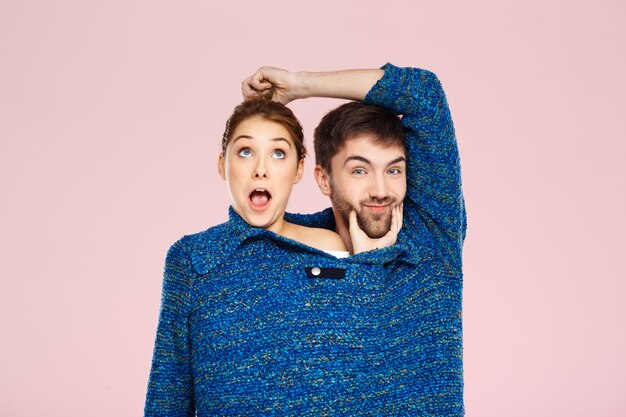 Junges schönes Paar in einem blauen Strickpullover posierend lächelnd, der Spaß über hellrosa Wand hat