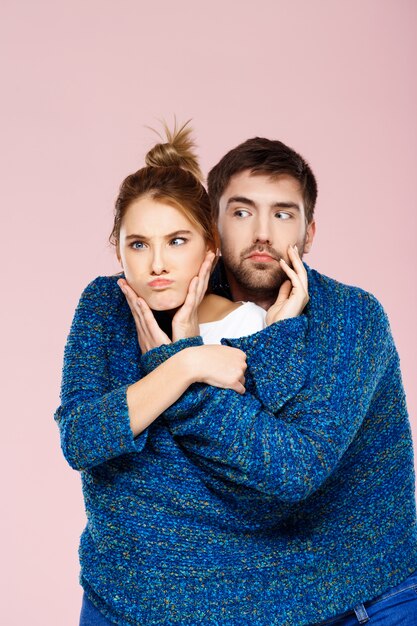 Junges schönes Paar in einem blauen Strickpullover posierend lächelnd, der Spaß über hellrosa Wand hat