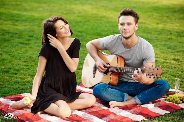 Junges schönes Paar lächelt, ruht, entspannt sich beim Picknick im Park. Mann, der Gitarre spielt.