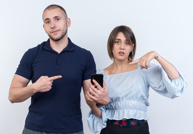 Junges schönes Paar, Mann und Frau, die einen Mann mit Smartphone suchen, der mit dem Zeigefinger auf seine verwirrte Freundin zeigt
