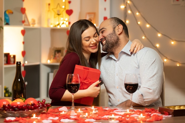 Junges schönes Paar Mann und Frau mit Geschenk, das am Tisch sitzt, verziert mit Kerzen und Rosenblättern glücklich in der Liebe, die internationalen Frauentag im dekorierten Wohnzimmer feiert