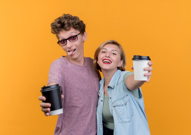 Junges schönes Paar Mann und Frau tragen Freizeitkleidung, die Kaffeetassen lächelnd über orange Wand stehend zeigt