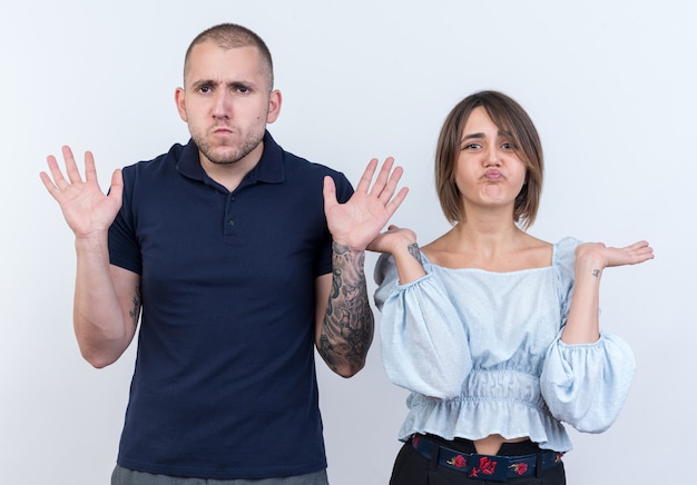 Kostenloses Foto junges schönes paar mann und frau verwirrt, die arme an den seiten auszubreiten und keine antwort zu haben, die über der weißen wand steht